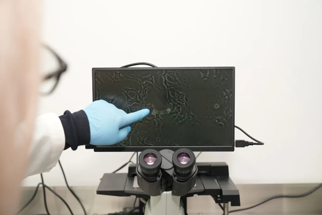 Gloved researcher pointing to cell activity on a microscope monitor in the Blast Institute lab